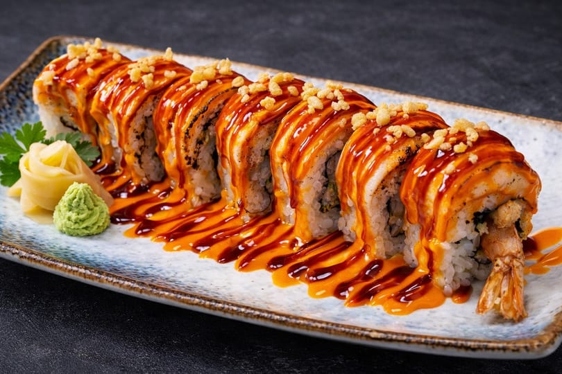 A sushi roll with shrimp, drizzled with orange and dark sauces, topped with crunchy bits.